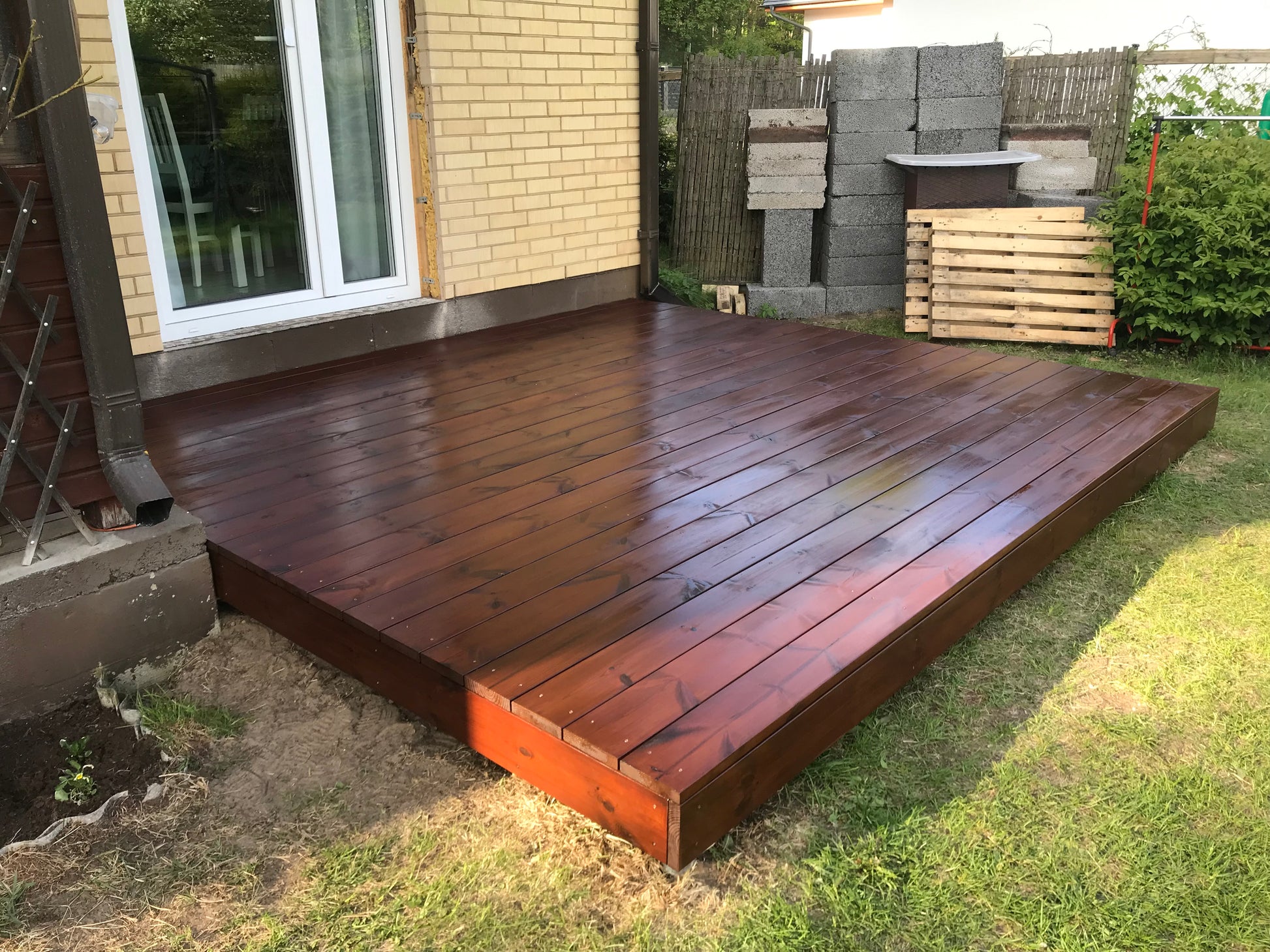 Wooden deck extending from a house with stacks of wooden planks in the background.