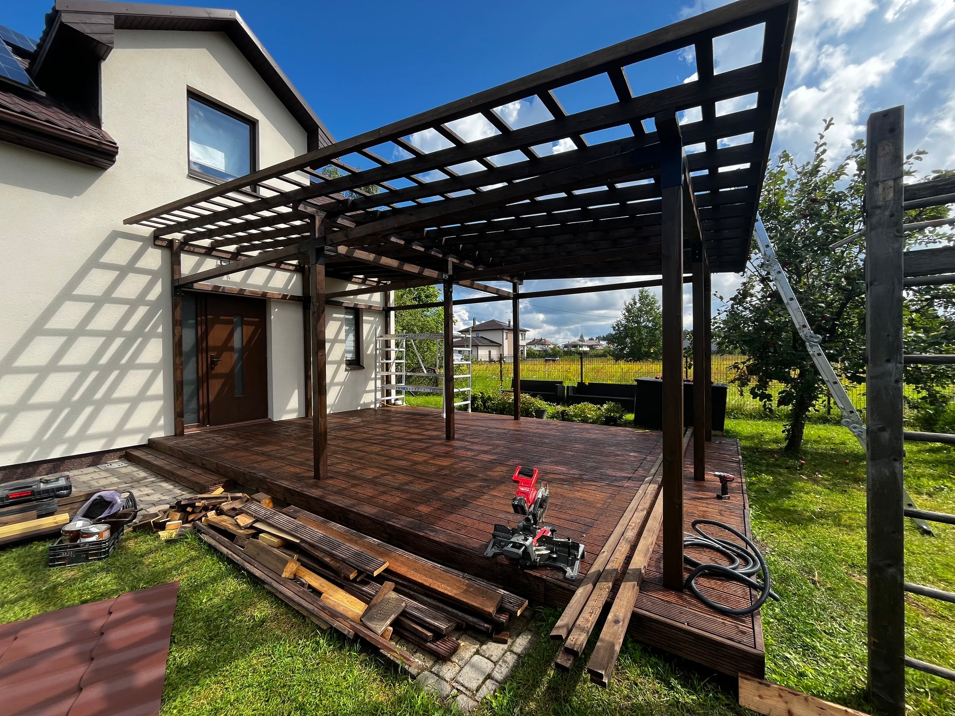 Wooden deck under a pergola with tools and materials on a grassy area.