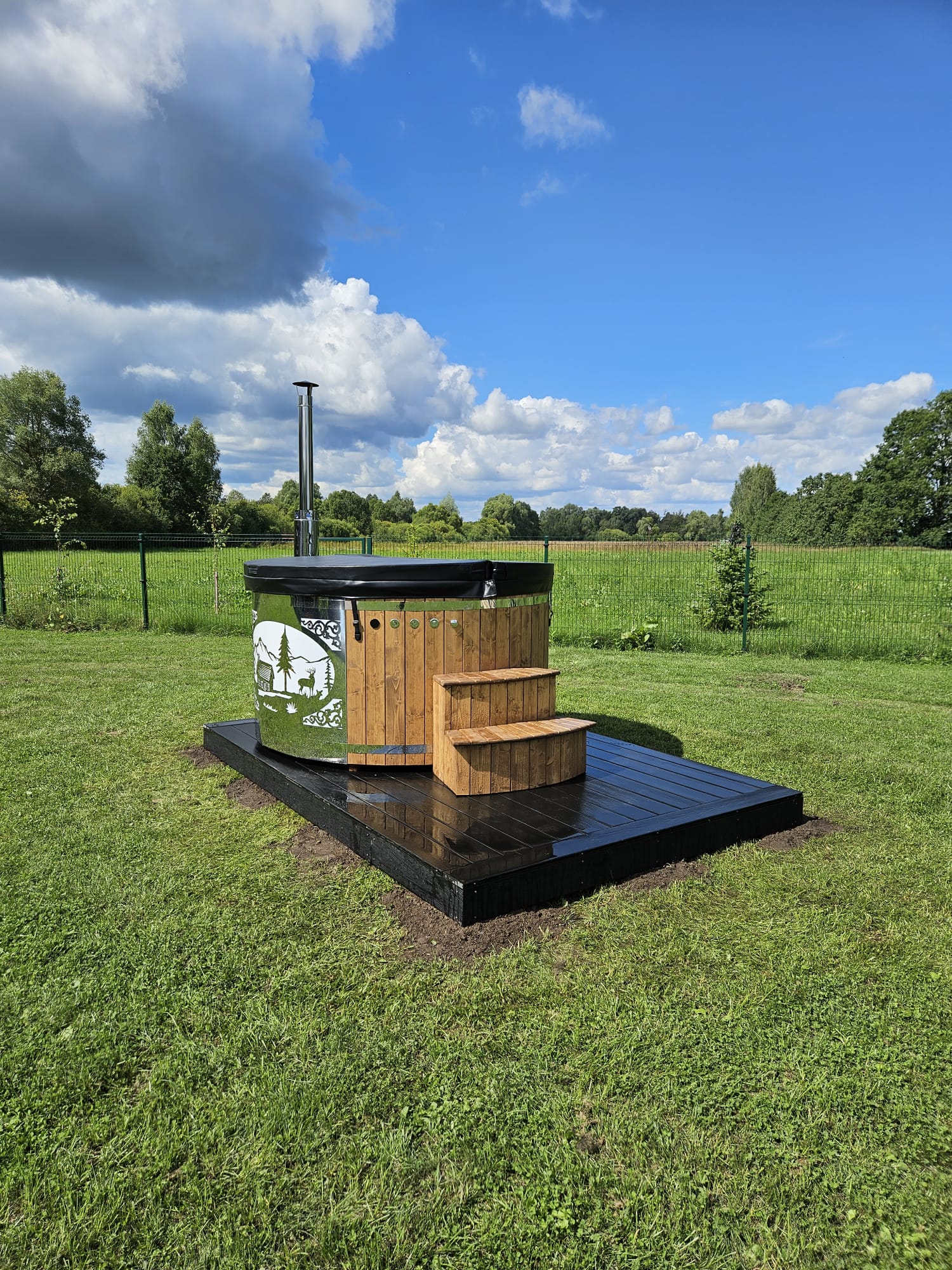 Outdoor hot tub with wooden steps in a grassy field under a blue sky with clouds.