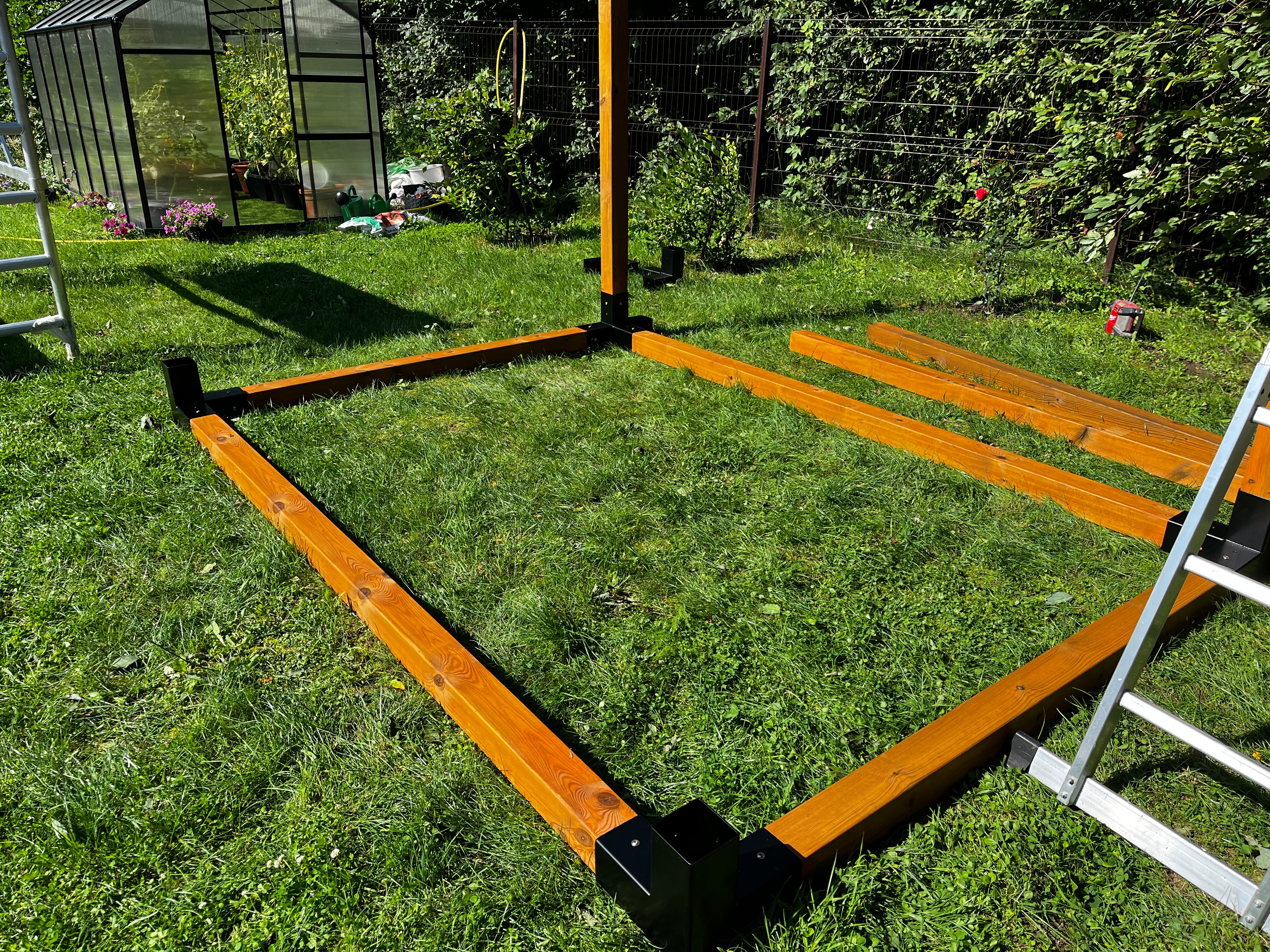 Wooden frame structure on grass with a ladder and garden area in the background
