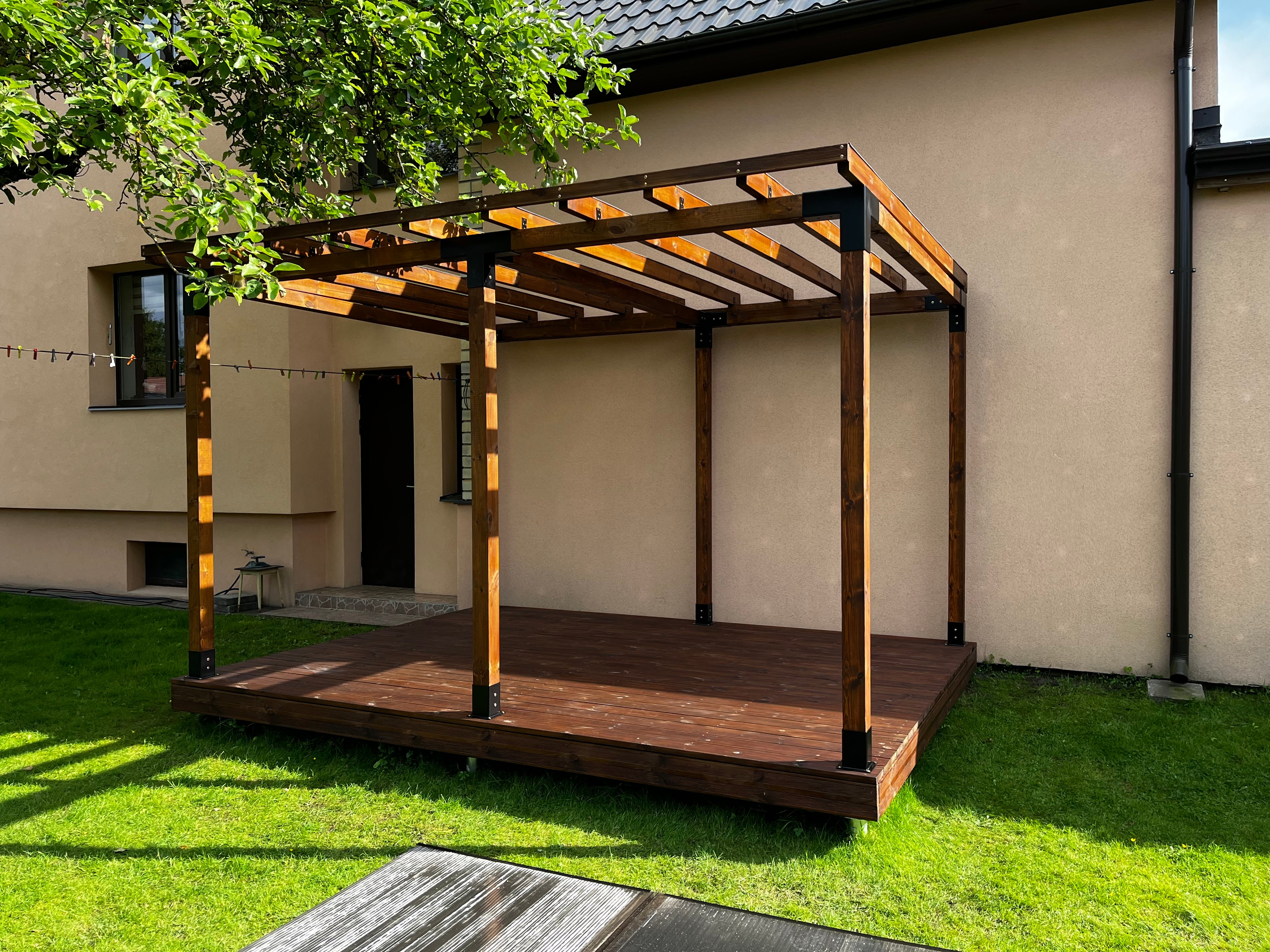 Wooden pergola attached to a house with green grass and a tree in the background