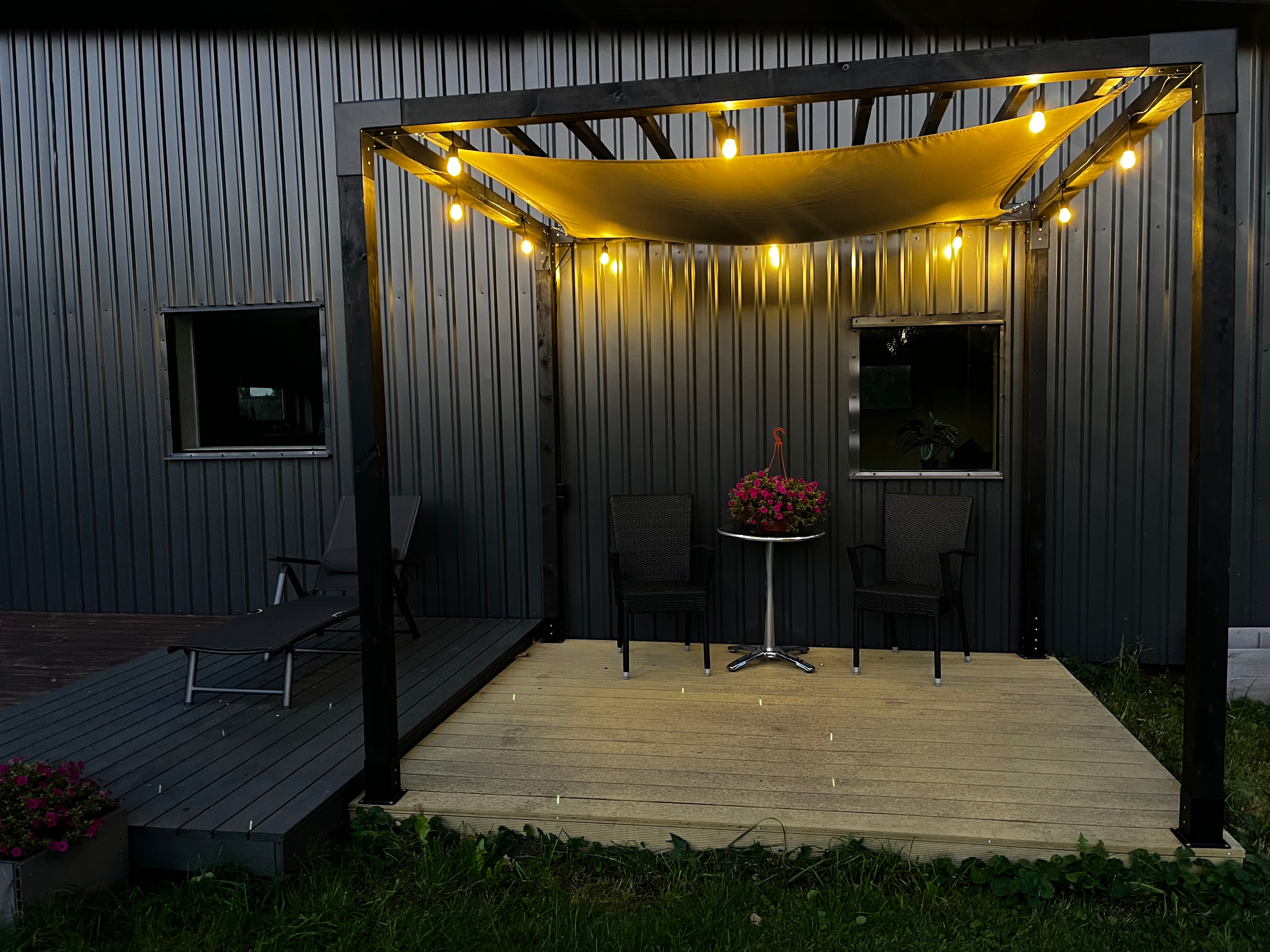 Outdoor patio area with a canopy, table, and chairs under string lights against a metal building.