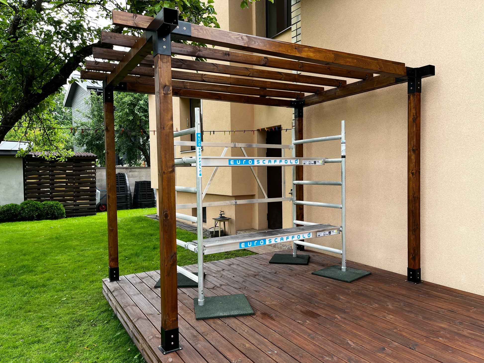 Wooden pergola on a wooden deck with a ladder leaning against a wall.