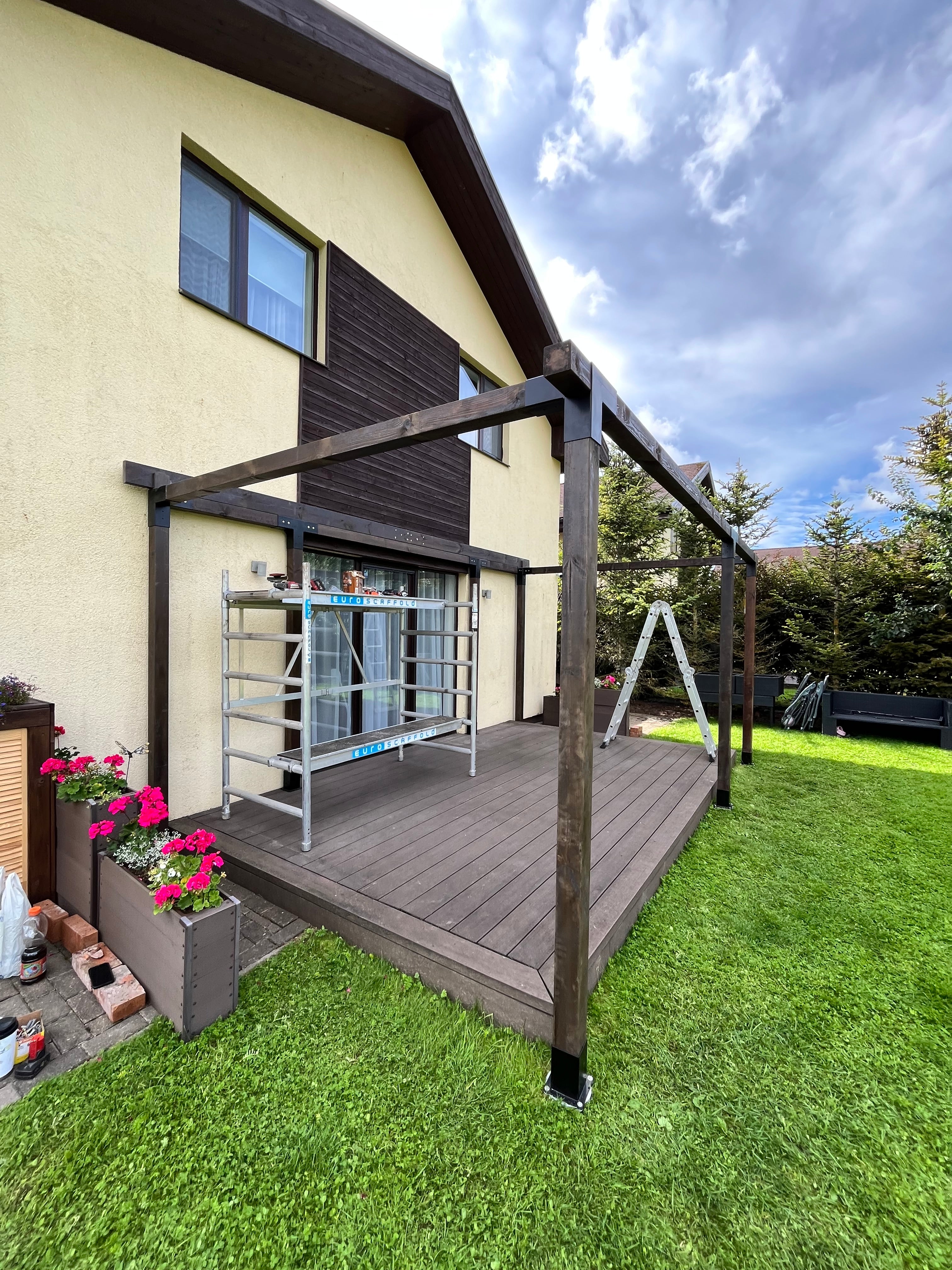 Wooden deck with canopy leading to a house with a green lawn and blue sky.