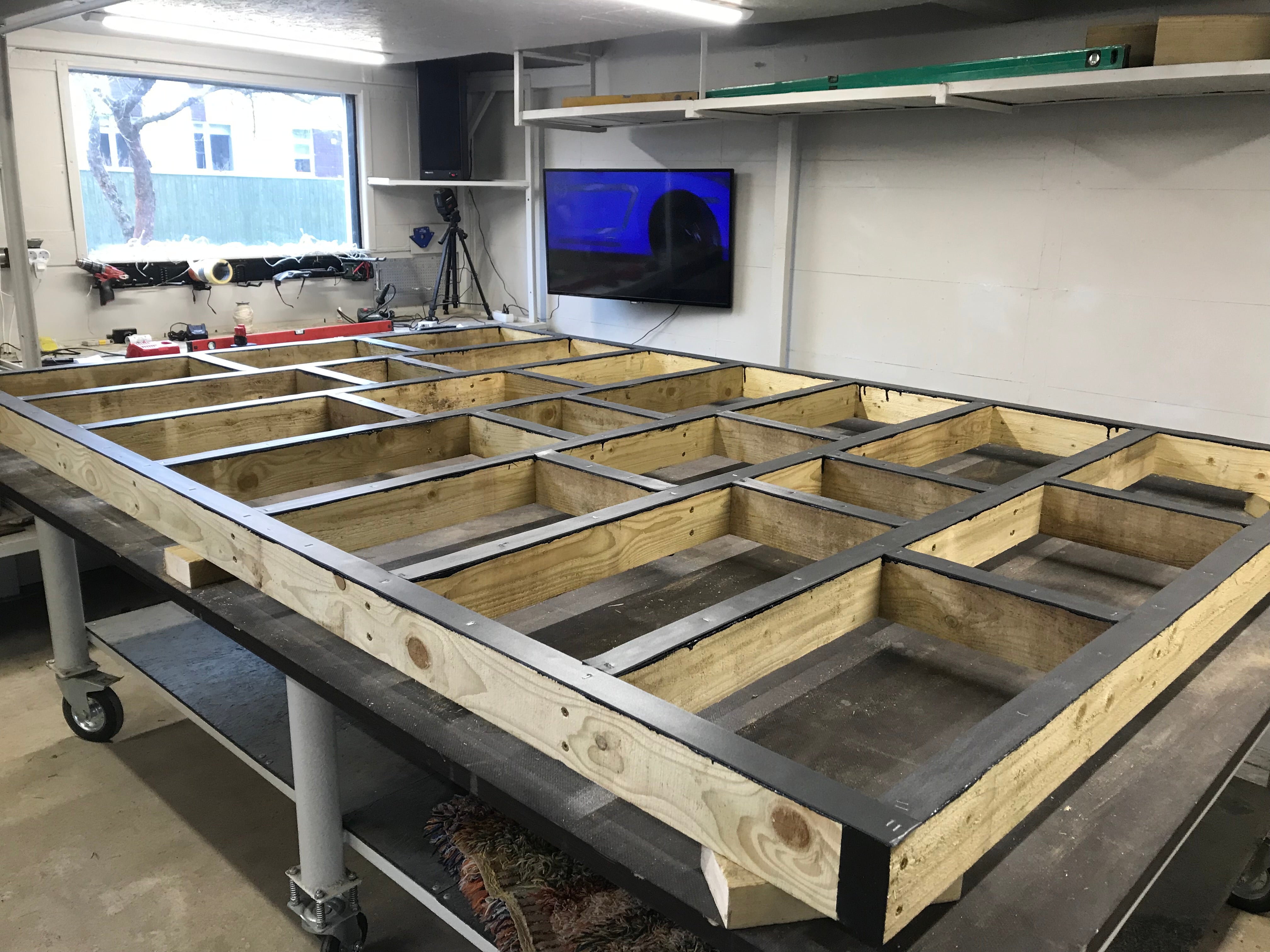 Wooden floor frame construction in a workshop setting with tools and equipment in the background.