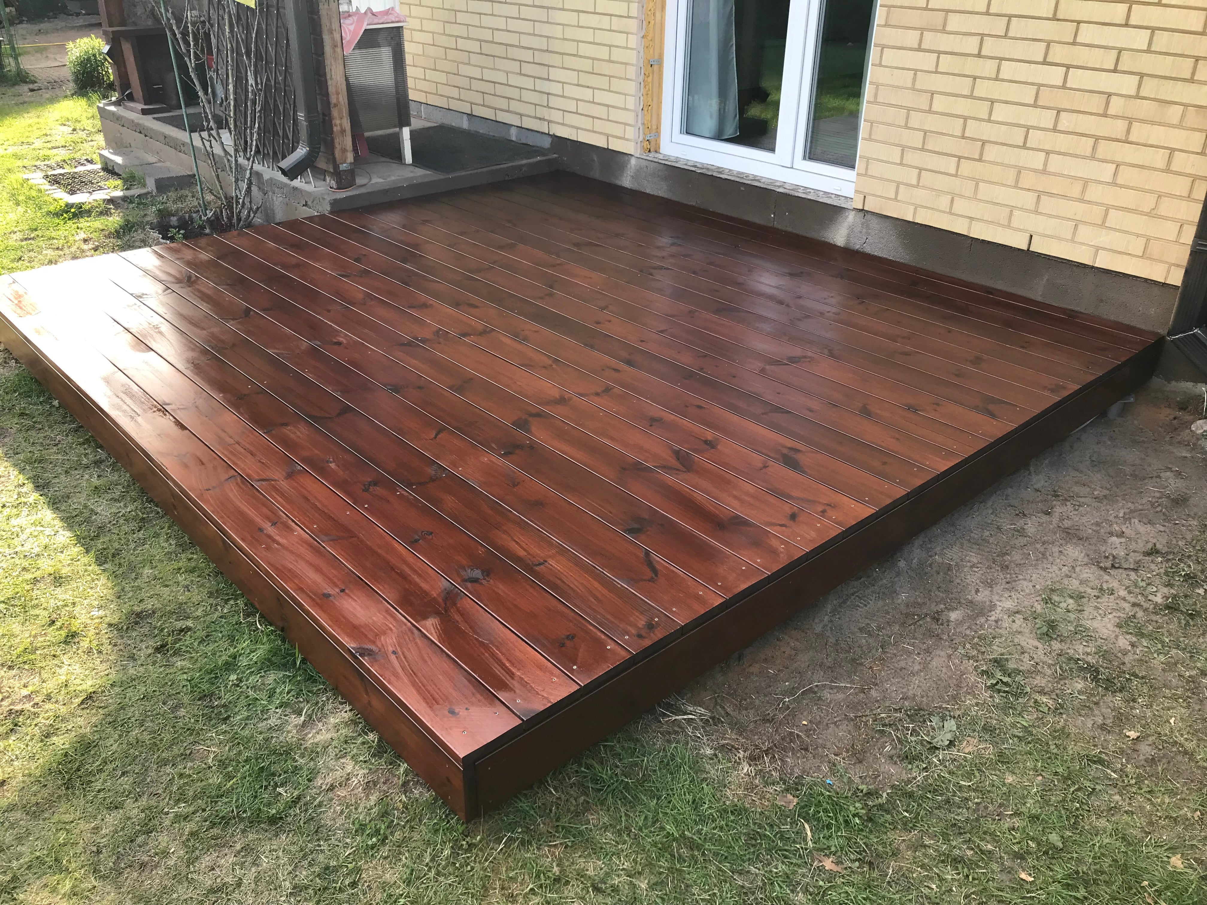 Wooden deck in front of a house with grass and a brick wall in the background.